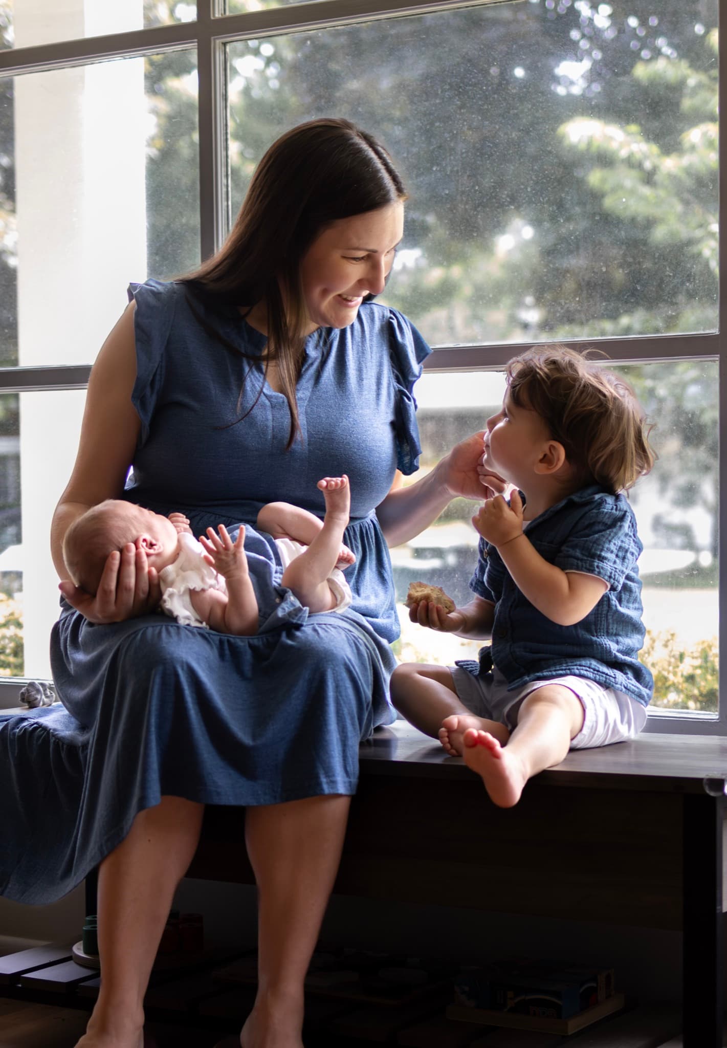 newborn photography session in Skokie home — mom holding newborn in natural window light with big brother beside her