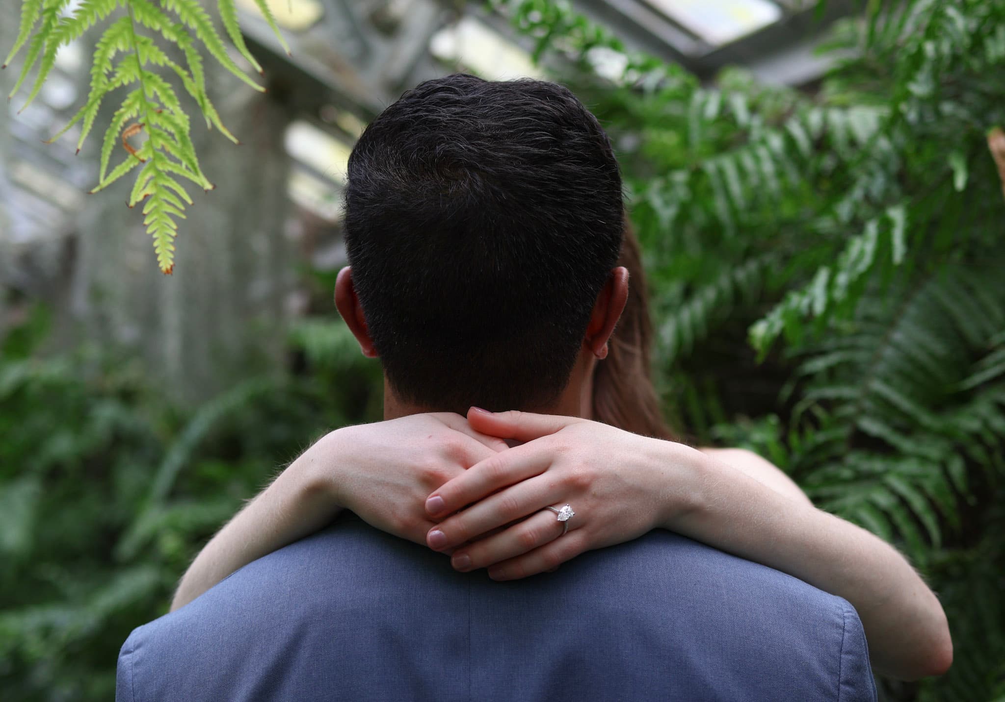 Bride-to-be hugging her fiance from behind with engagement ring visible in the greenhouse