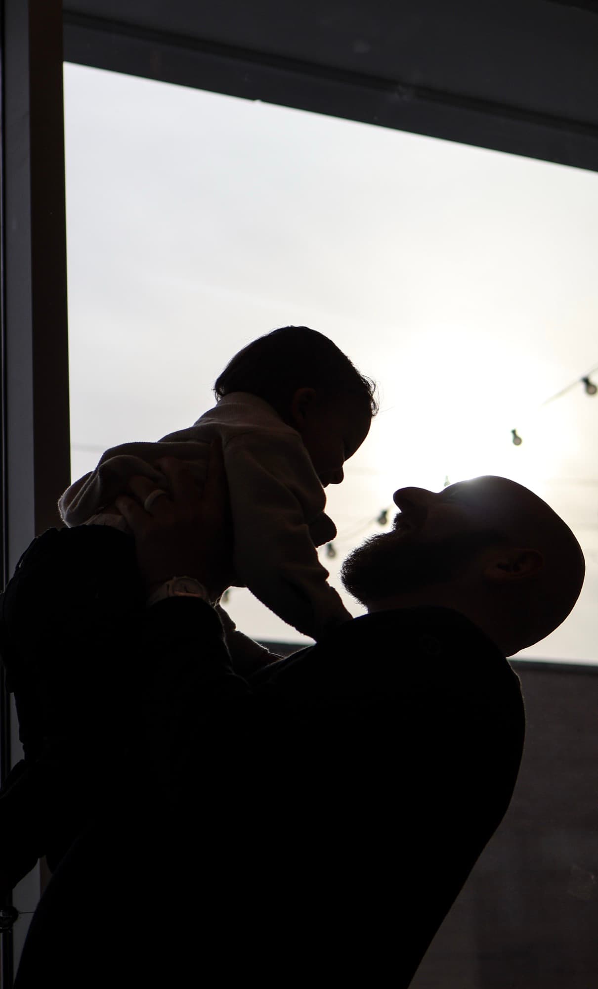 Father holding baby up in window silhouette — artistic family portrait