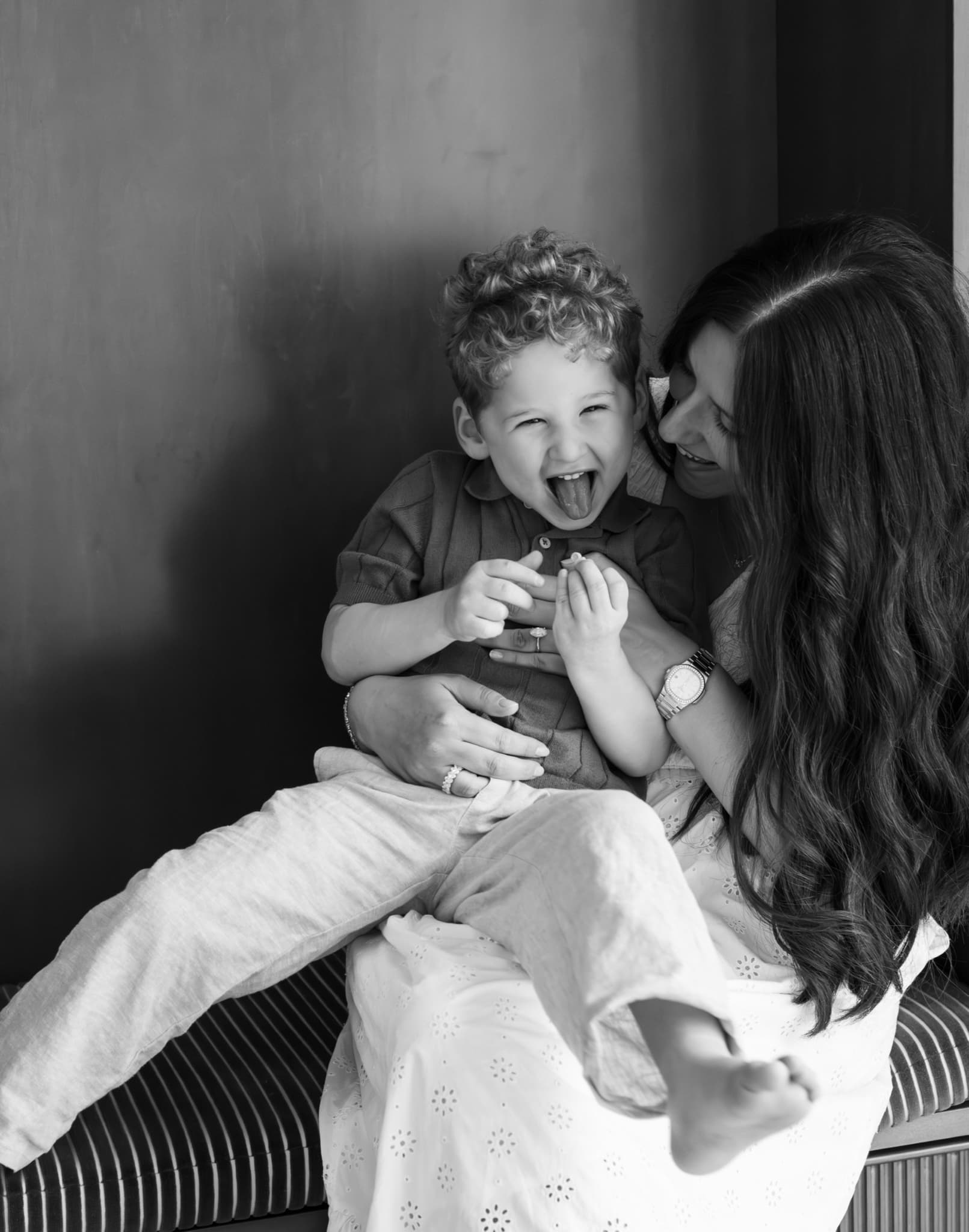 Black and white photo of laughing boy sticking his tongue out on mom's lap — playful family photography