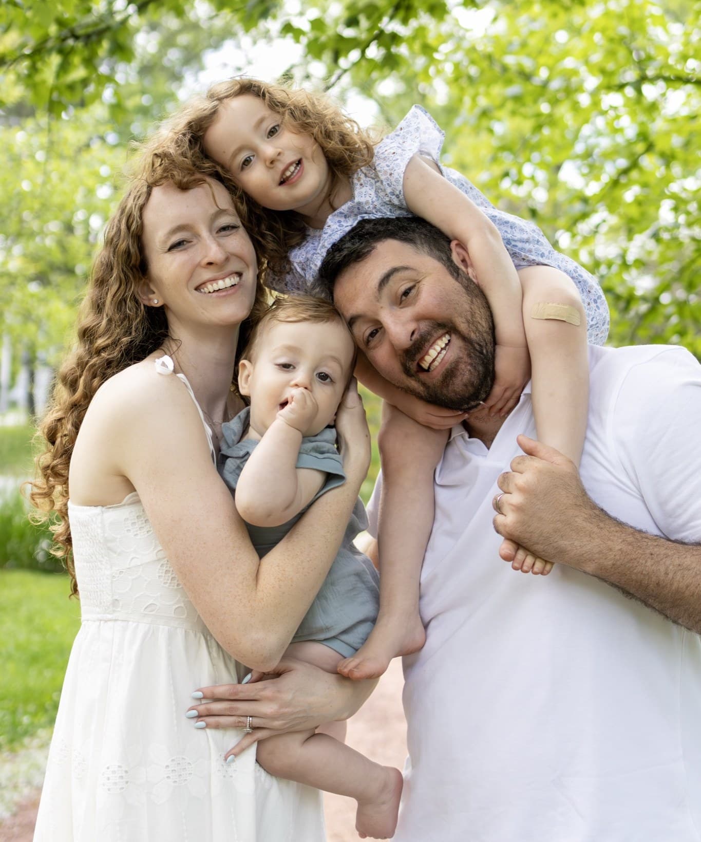 Family of four smiling under a green tree canopy — daughter on dad's shoulders, baby on mom's hip