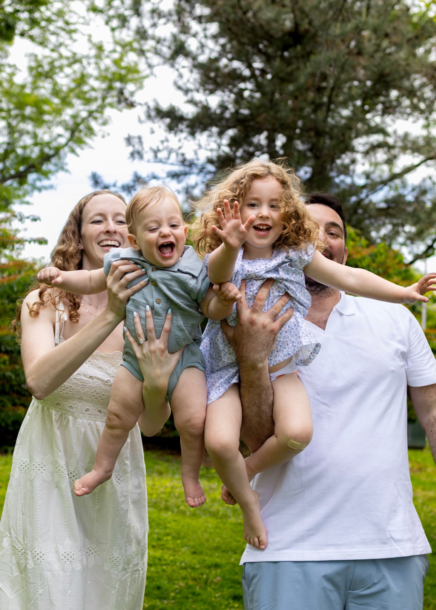 Parents lifting their laughing toddler and daughter toward the camera in the park — playful family photography