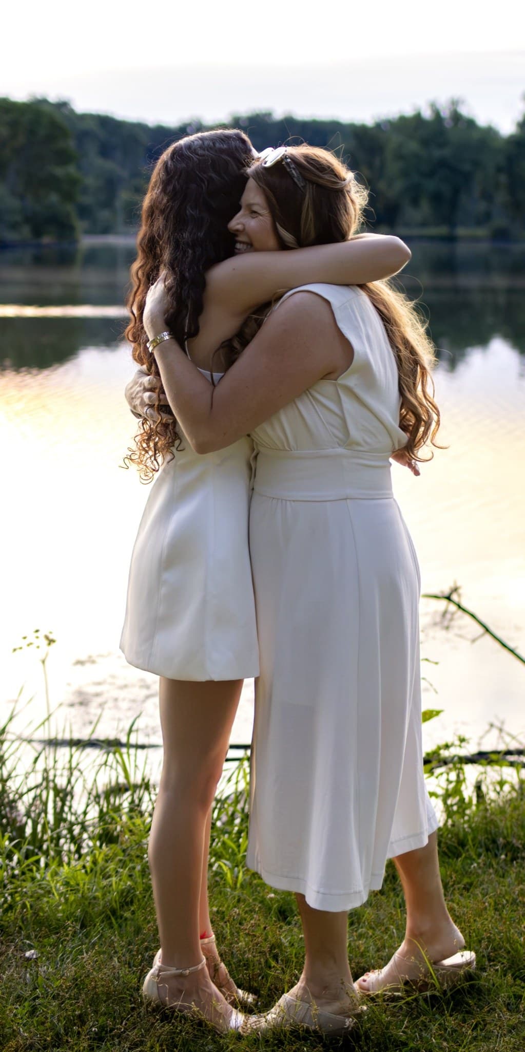 Two women hugging lakeside in white dresses at golden hour — candid photography