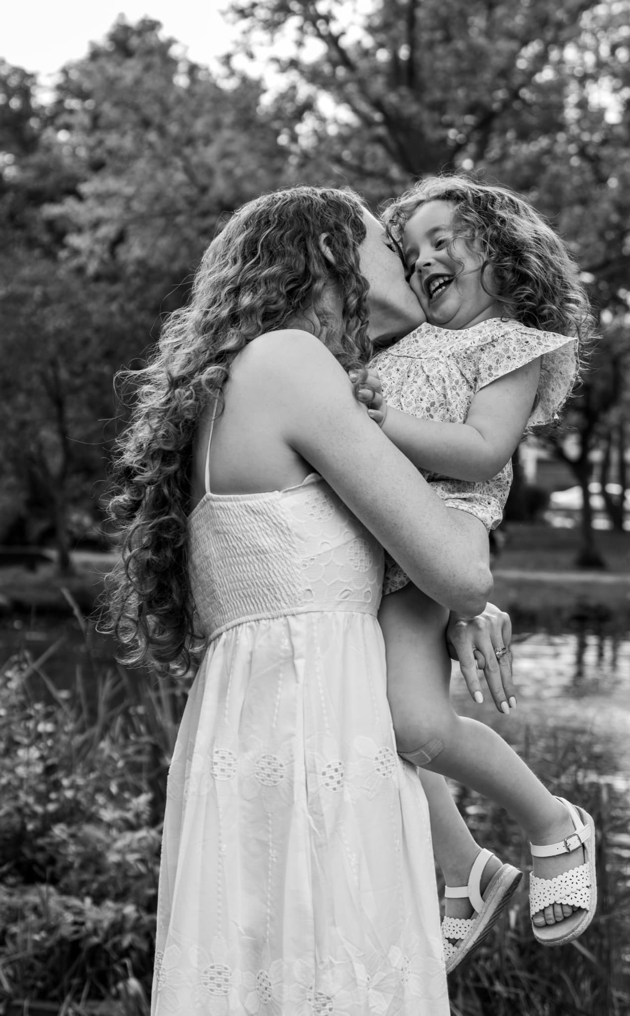 Black and white photo of mom lifting and kissing her laughing curly-haired daughter by the pond — family photography
