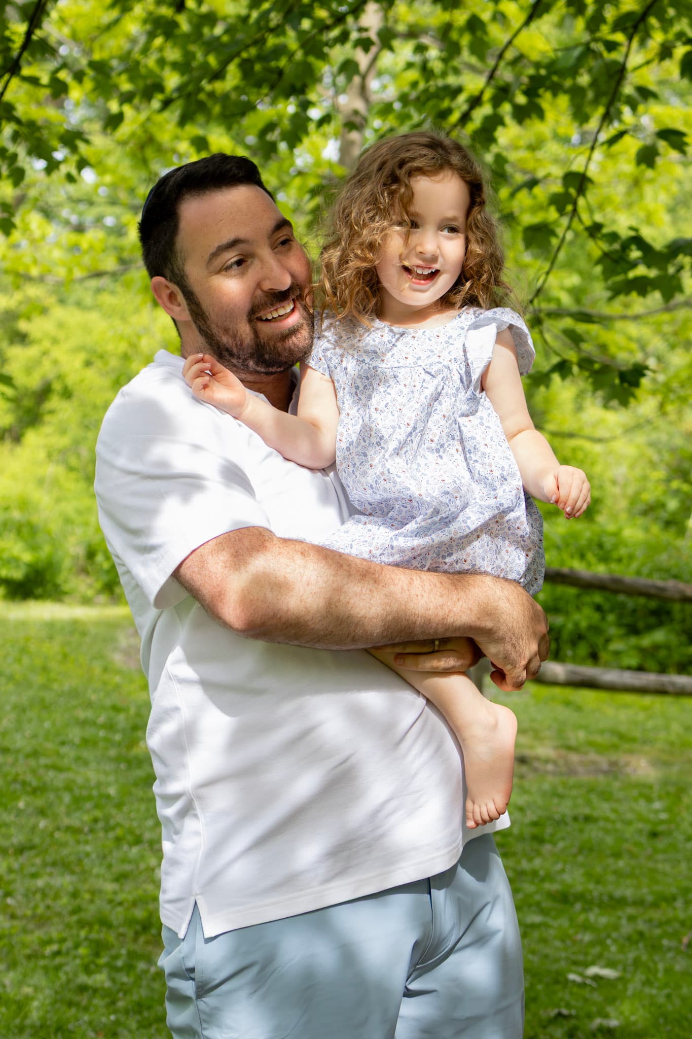 Dad in white tee holding smiling daughter in floral dress during an outdoor family session in Wilmette