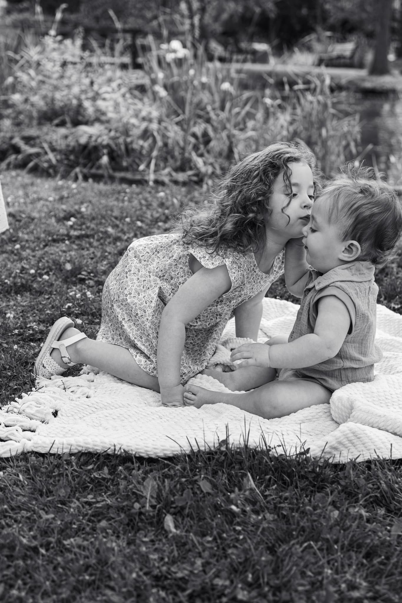 Black and white moment of big sister kissing baby brother on a blanket outdoors at Keay Nature Center