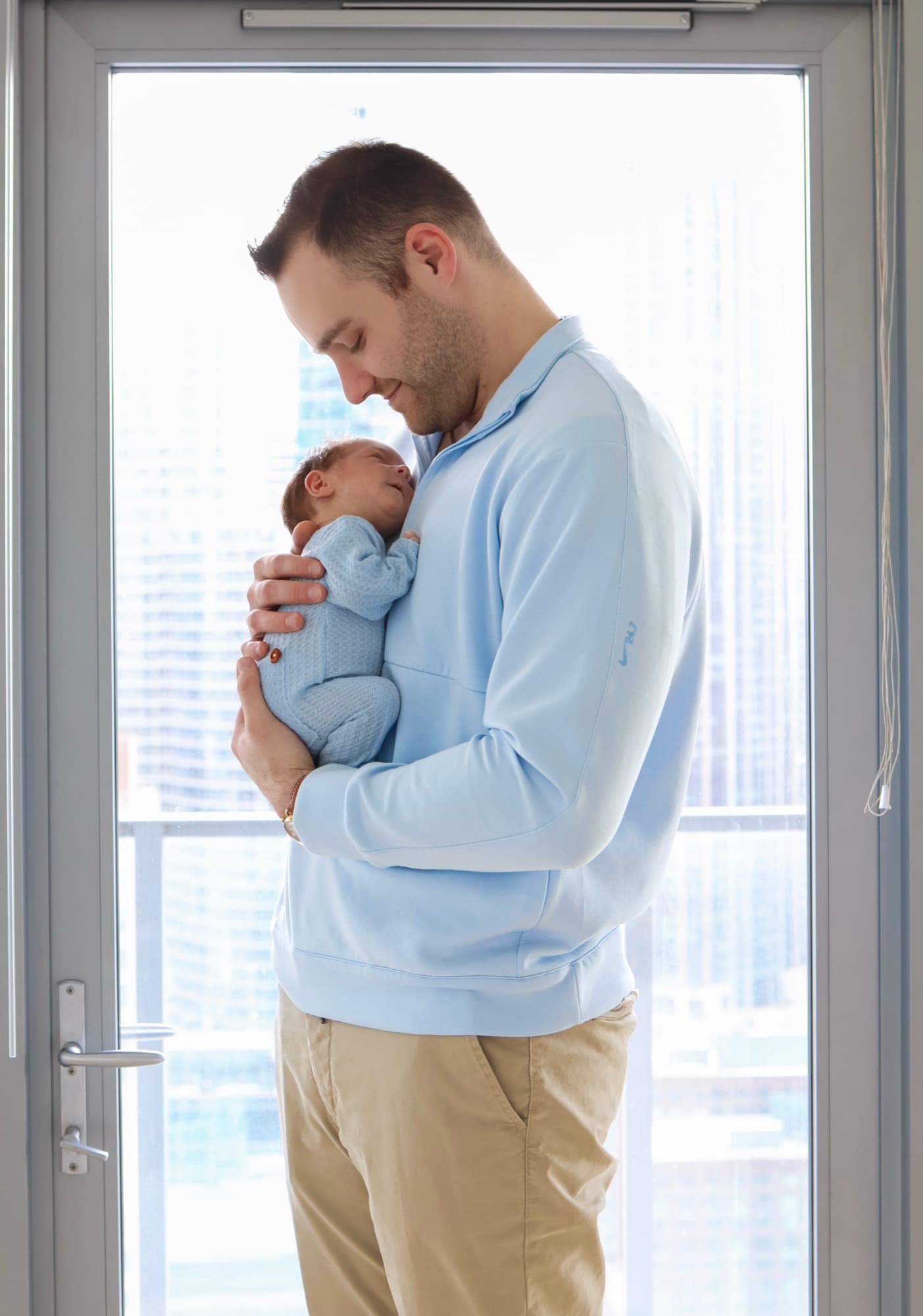 Father holding newborn by window with city skyline — downtown newborn photography