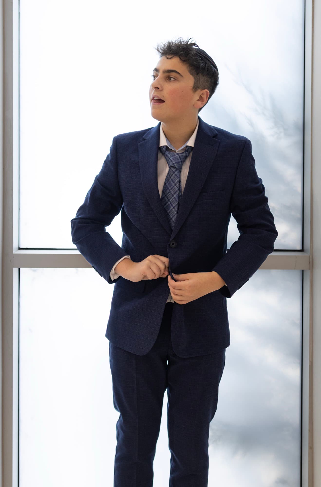 Bar Mitzvah boy buttoning suit by window — editorial getting-ready portrait