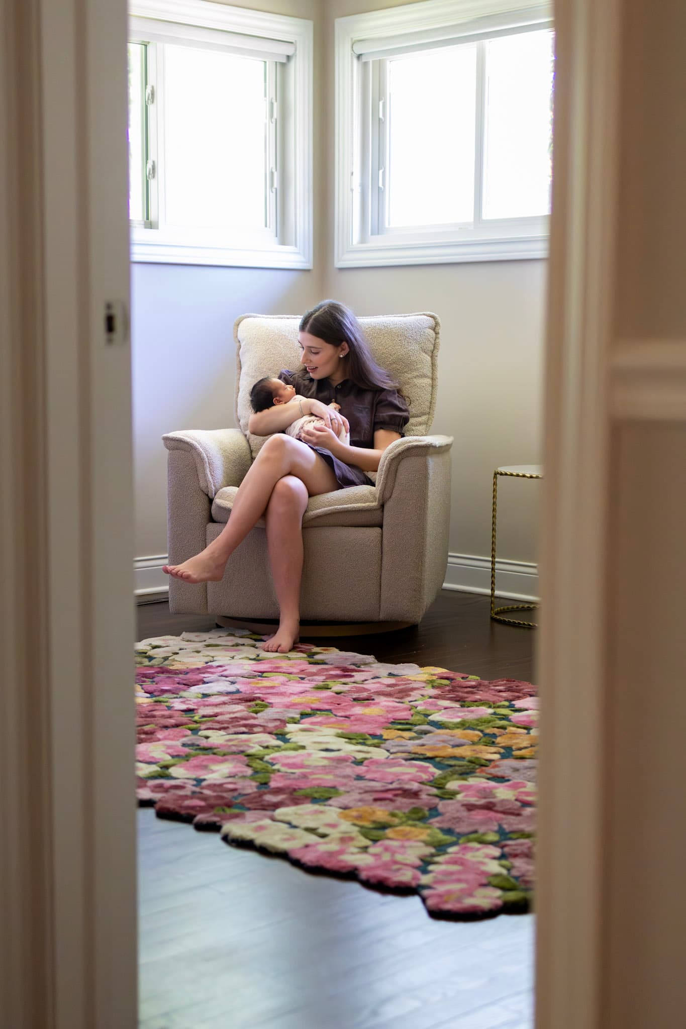 In-home newborn lifestyle moment — mom holding baby girl in nursery glider above a floral rug in Skokie