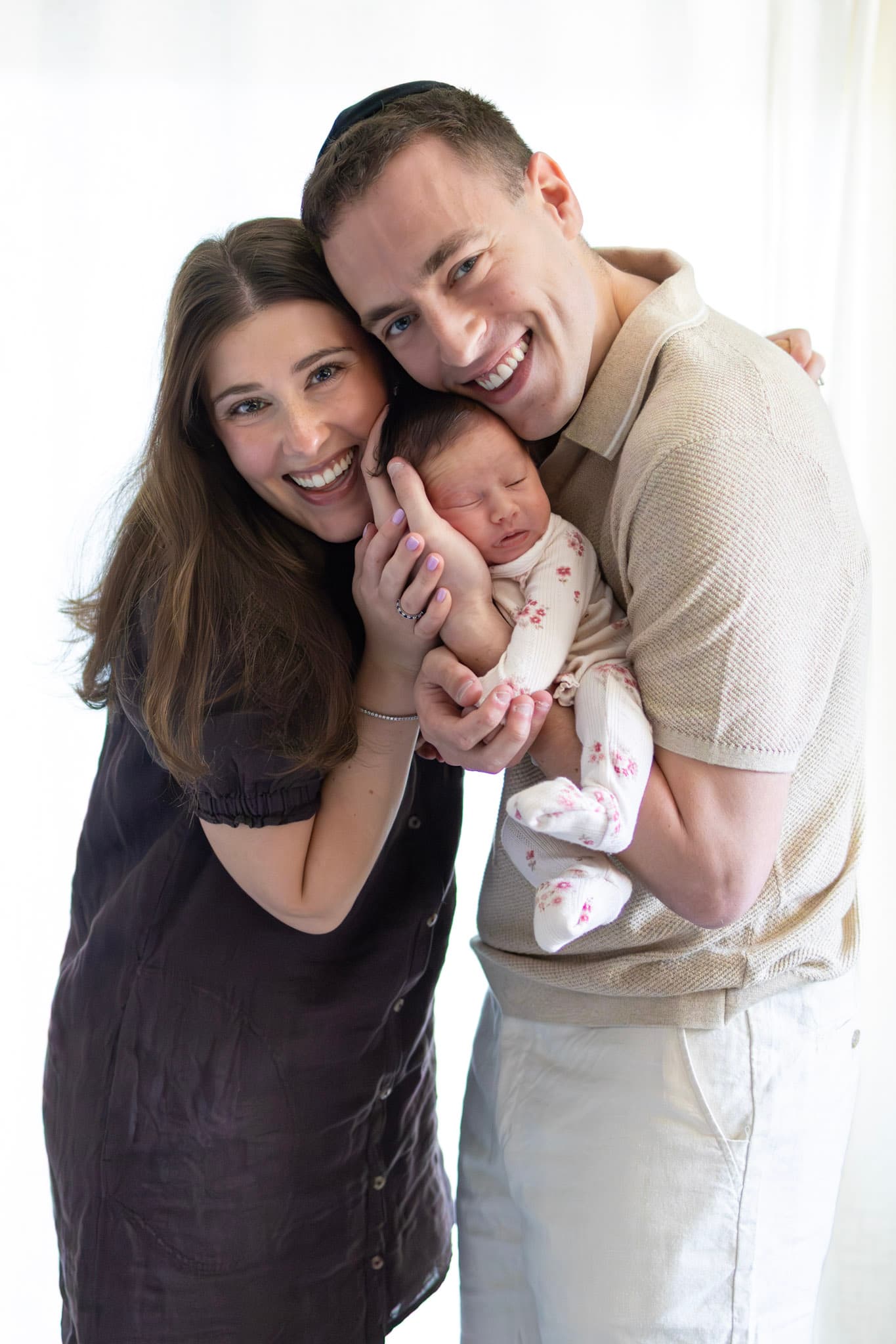 Skokie newborn family portrait — young parents cheek to cheek smiling with baby girl in floral romper