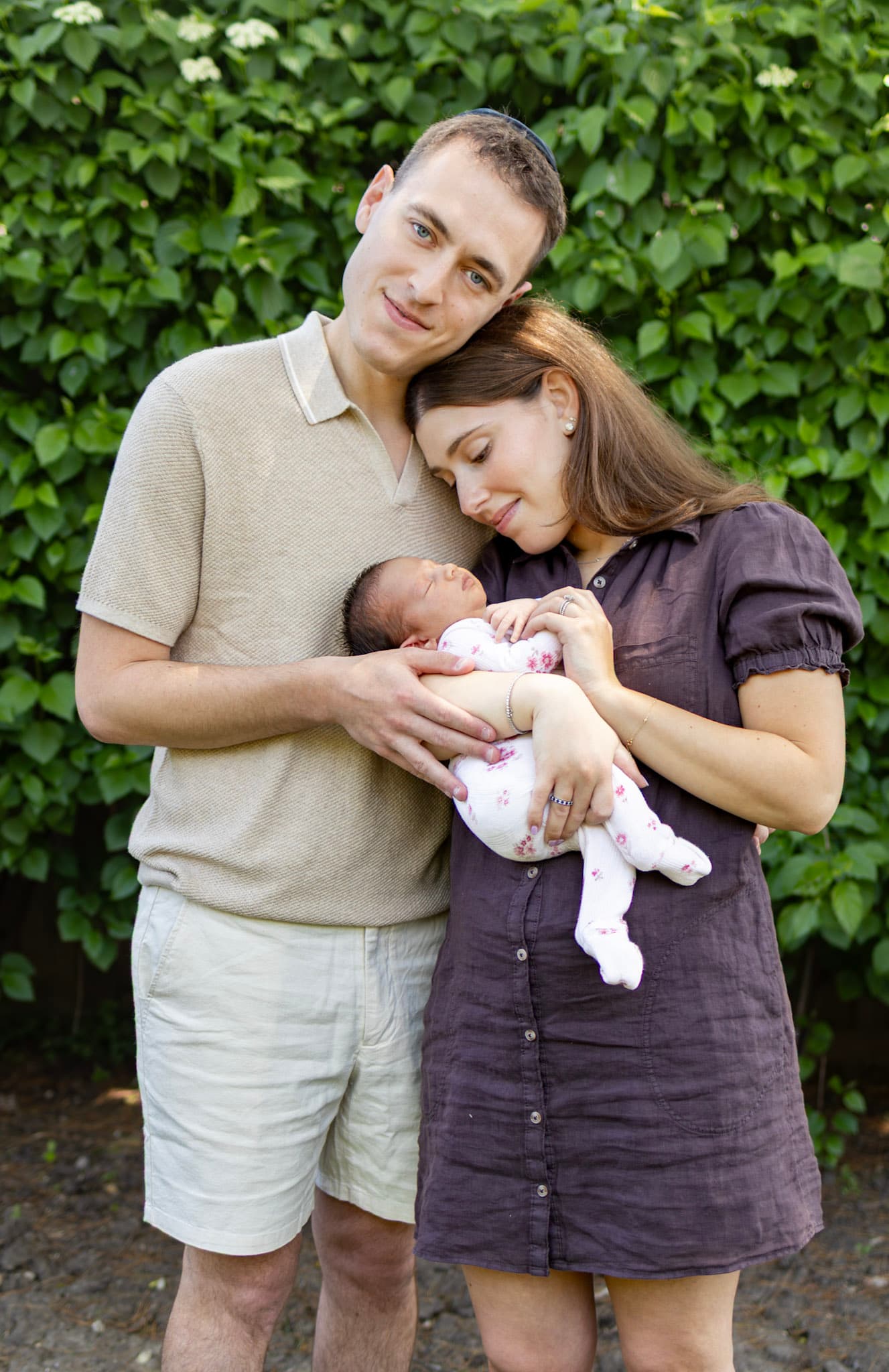 Outdoor newborn session in Skokie — parents holding baby girl against green ivy hedge in summer light