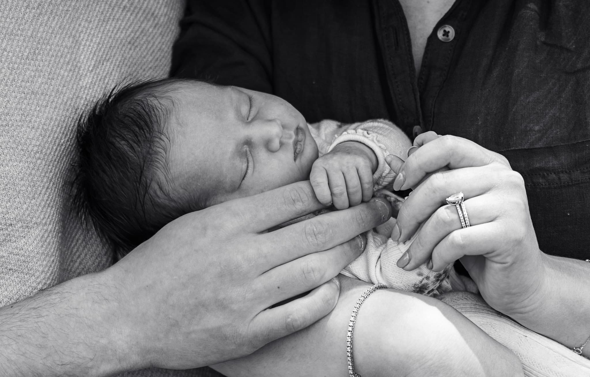 Black and white detail photo — parents' hands and wedding rings cradling newborn baby girl